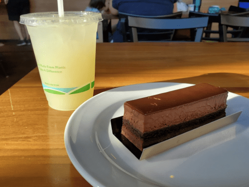 A slice of chocolate cake on a plate next to a cup of lemonade on a wooden table.