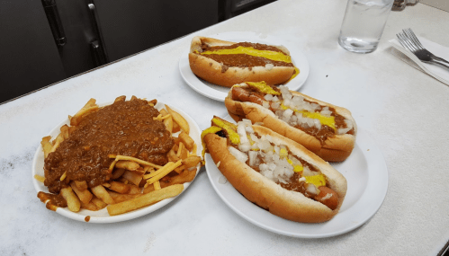 A plate of chili cheese fries next to two hot dogs topped with mustard and onions on a white table.
