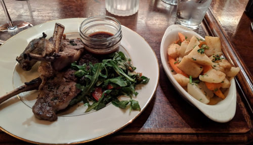 A plate with grilled lamb chops, a small jar of sauce, arugula salad, and a side of roasted root vegetables.