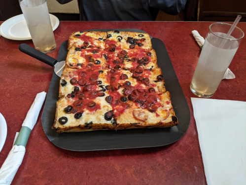 A rectangular pizza topped with pepperoni and black olives on a black serving tray, with drinks in the background.
