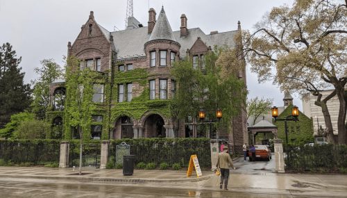 A large, ivy-covered mansion with turrets, surrounded by trees, on a rainy day. A person walks by on the wet sidewalk.