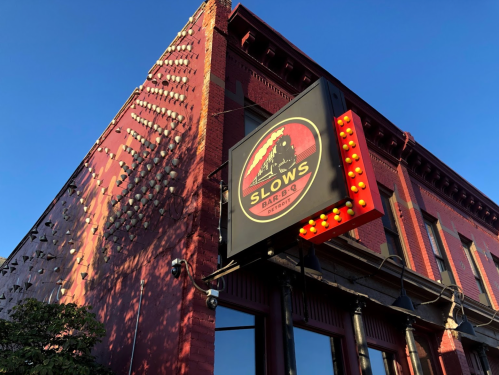 A red brick building with a sign for "Slows Bar BQ" featuring bright lights against a clear blue sky.