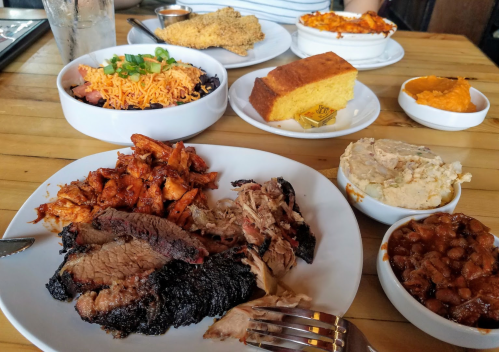 A table spread with various dishes, including smoked meats, cornbread, beans, and sides, on a wooden surface.