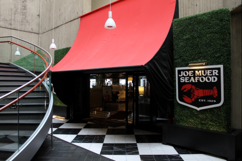Entrance to Joe Muer Seafood, featuring a red awning, greenery, and a logo, with a spiral staircase nearby.