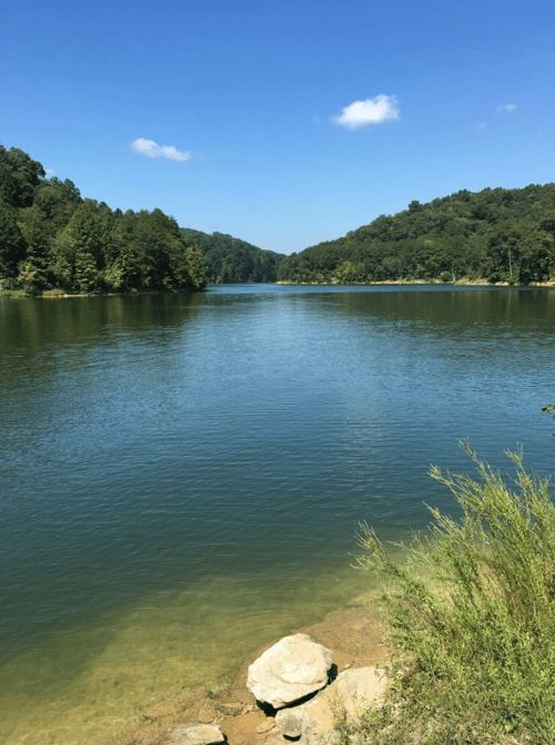 A serene lake surrounded by green hills under a clear blue sky, with a rocky shore in the foreground.