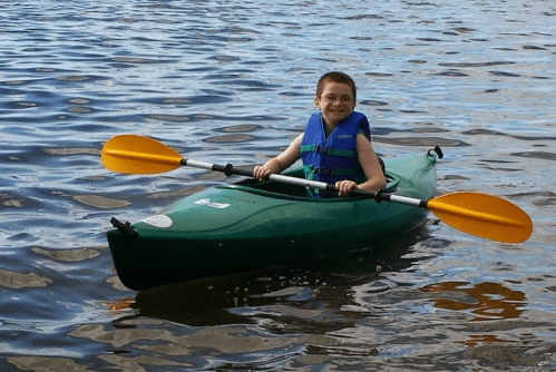 A smiling child in a green kayak with orange paddles, enjoying a sunny day on the water.