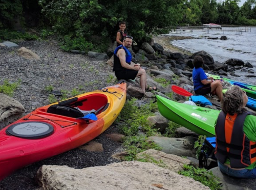 A group of people in life jackets by the water, with colorful kayaks on the shore and greenery in the background.