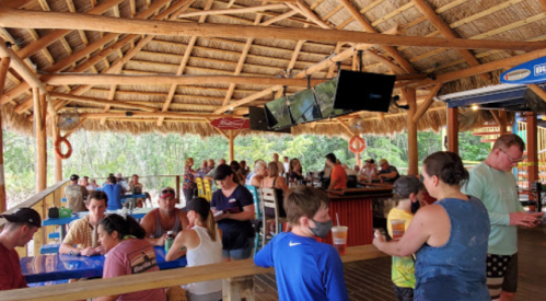 A lively outdoor bar with thatched roof, filled with people enjoying drinks and conversation in a tropical setting.