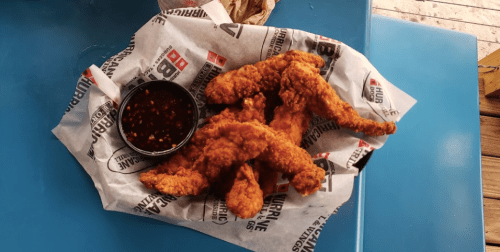 A plate of crispy fried chicken tenders served with a side of dipping sauce on a paper-lined tray.