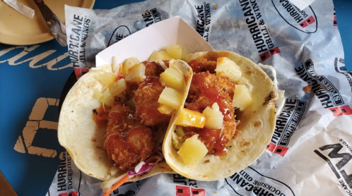 Two tacos filled with crispy chicken, pineapple chunks, and sauce, served on a branded paper.