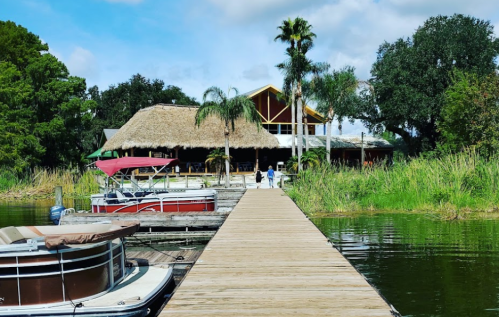 A wooden dock leads to a thatched-roof building surrounded by greenery and boats on the water.