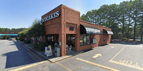 A brick building with a sign reading "Cococakes," surrounded by trees and a parking lot on a sunny day.