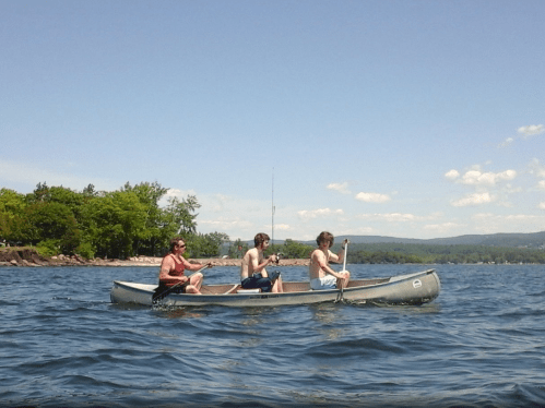 Three people paddle a canoe on a calm lake, surrounded by trees and a clear blue sky.