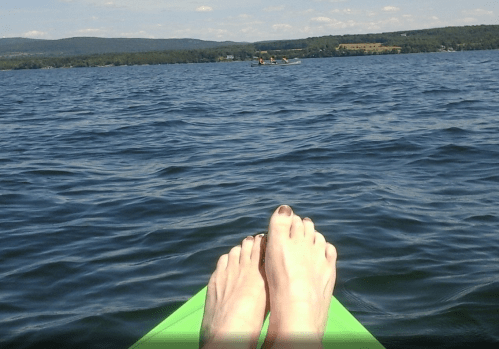 A pair of bare feet resting on the edge of a kayak, with a calm lake and distant hills in the background.