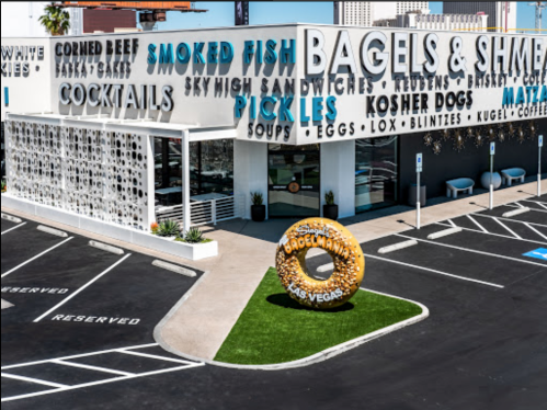 Exterior of a bagel shop featuring a large donut sculpture, with signage for various food items and a parking lot.