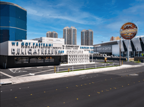 A modern deli and shopping center with large signage, set against a city skyline and clear blue sky.