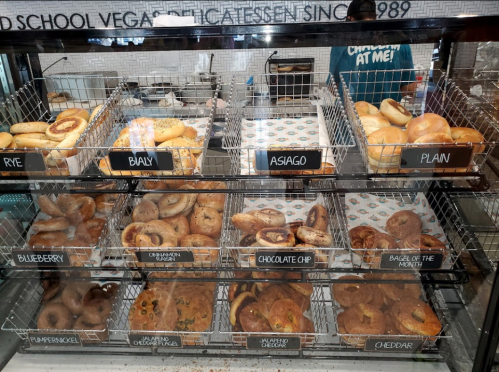 A display case filled with various bagels, including rye, bialy, asiago, plain, blueberry, and chocolate chip.
