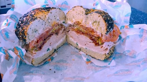 A halved bagel sandwich filled with turkey, lettuce, tomato, and crispy bacon, wrapped in branded paper.