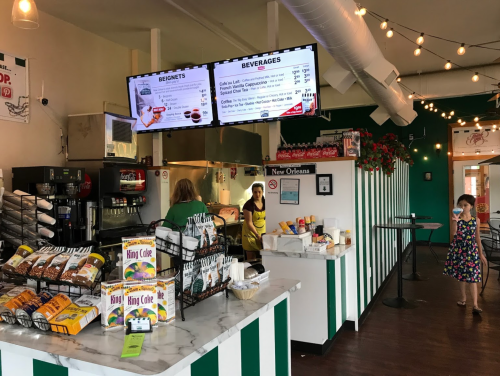 A colorful café interior with a menu display, a counter, and a customer in a dress, while staff prepares orders.