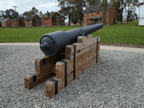 A historic cannon mounted on a wooden carriage, set on gravel with old brick buildings in the background.