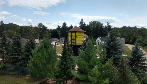 A tall yellow structure on stilts surrounded by green trees and houses under a blue sky with scattered clouds.