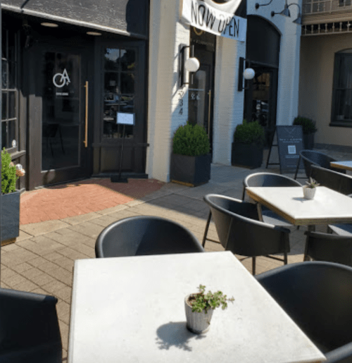 Outdoor café scene with tables, potted plants, and a sign reading "NOW OPEN" on a sunny day.