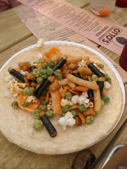 A plate with a tortilla topped with popcorn, green peas, and various snack sticks.