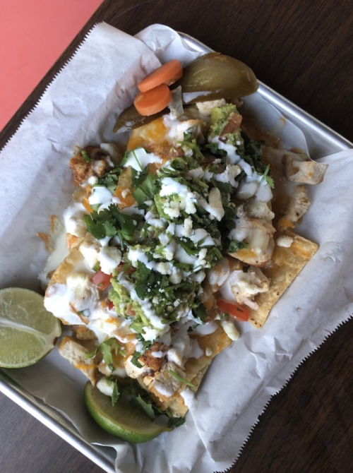 A tray of nachos topped with cheese, guacamole, sour cream, diced tomatoes, onions, and garnished with lime and pickled vegetables.