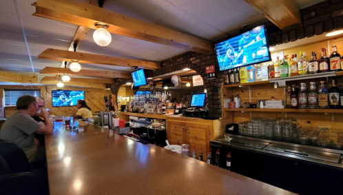 A cozy bar with wooden decor, two patrons at the counter, and multiple TVs displaying sports. Bottles line the shelves.