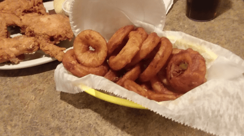 A basket of golden-brown onion rings next to a plate of fried chicken.