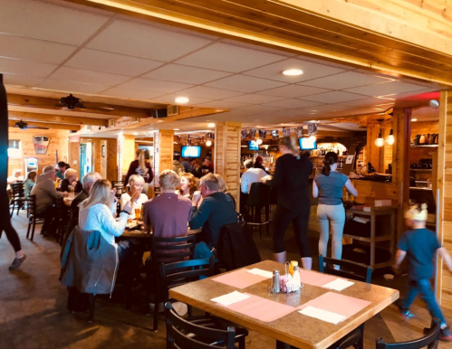 A busy restaurant interior with diners at tables, a bar area, and staff serving customers in a warm, wooden setting.