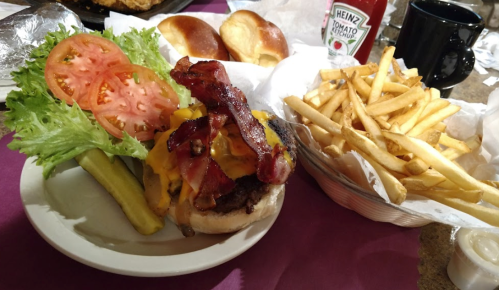 A plate with a cheeseburger topped with bacon, lettuce, tomato, pickles, and a side of fries. Ketchup and a coffee cup are nearby.