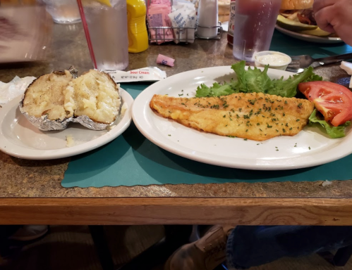 A plate with a fried fish fillet, lettuce, and tomato, alongside a baked potato on a restaurant table.