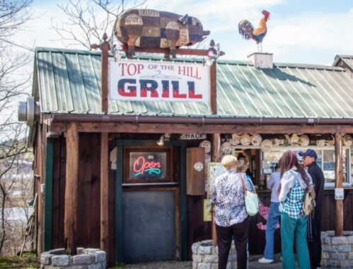 A rustic restaurant named "Top of the Hill Grill" with a rooster sign and people waiting outside.
