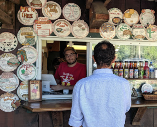A man orders at a food stand with colorful signs and menus displayed on the wall behind the counter.