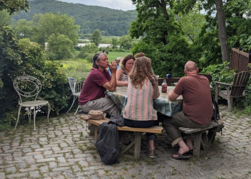 A group of people sitting around a table outdoors, surrounded by greenery and a scenic view.