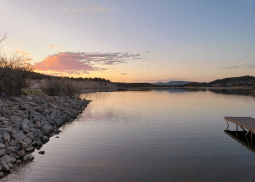 A serene lake at sunset, with rocky shores and a calm water surface reflecting the colorful sky.