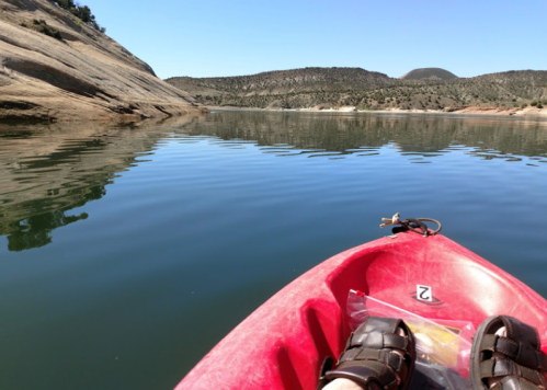 A view from a red kayak on calm water, with rocky shores and hills in the background under a clear blue sky.
