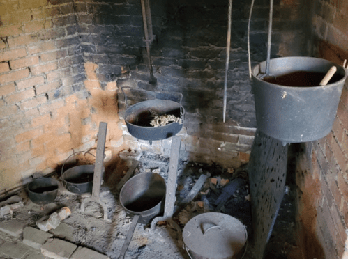 An old brick fireplace with several cast iron pots and a cooking pot hanging above, surrounded by soot and debris.