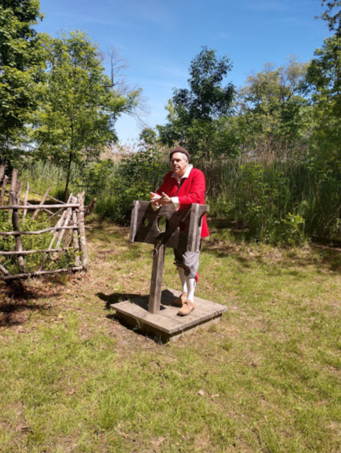 A person in historical attire stands by a wooden stocks in a grassy area with trees in the background.