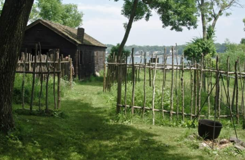 A rustic wooden cabin surrounded by a stick fence, with lush greenery and a view of a waterway in the background.
