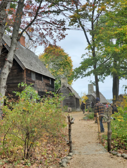 A path leads through a historic village with wooden houses and trees, set against a cloudy sky.