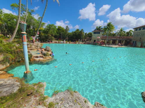 A clear turquoise pool surrounded by palm trees and rocky edges, with people swimming and relaxing under a blue sky.