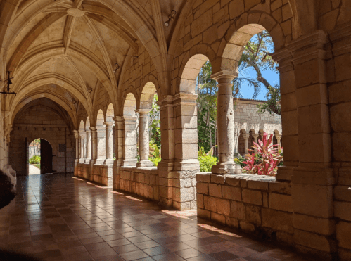 A stone corridor with arches and sunlight, featuring lush greenery and colorful plants outside.