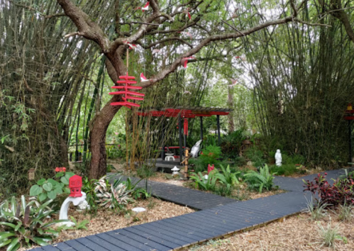 A serene garden with bamboo, a wooden path, red decorations, and a shaded seating area surrounded by lush greenery.