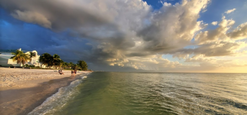 A serene beach scene with gentle waves, palm trees, and dramatic clouds under a colorful sky at sunset.