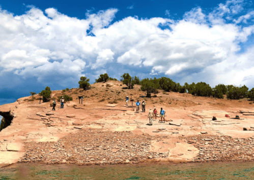 A rocky shoreline with people exploring under a partly cloudy sky and green trees in the background.