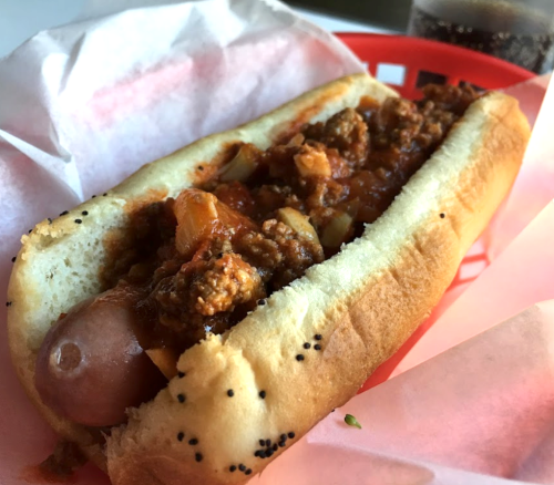 A hot dog in a poppy seed bun topped with chili and onions, served in a red basket with pink paper.
