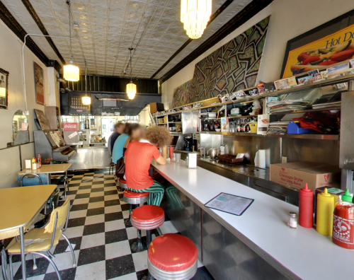 A retro diner interior with a long counter, red stools, and checkered floor, featuring patrons and colorful decor.