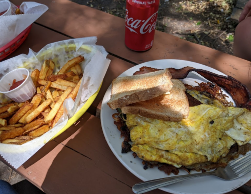A plate with eggs, bacon, toast, and fries, alongside a can of Coca-Cola on a wooden table.
